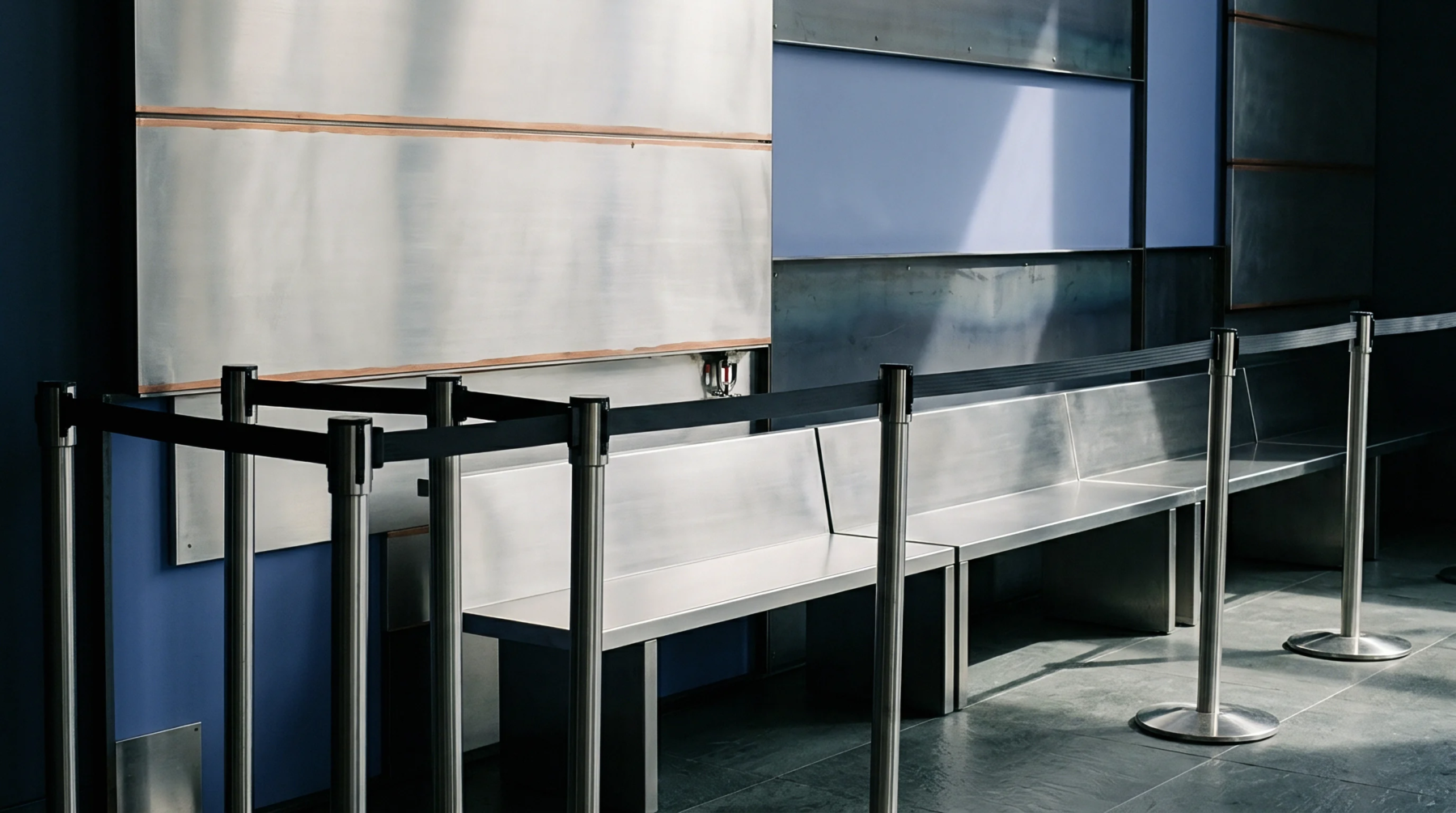 Stainless steel queue barriers with black retractable belts organize visitor flow near modular metal wall panels and durable benches in an industrial or large-scale operational facility.