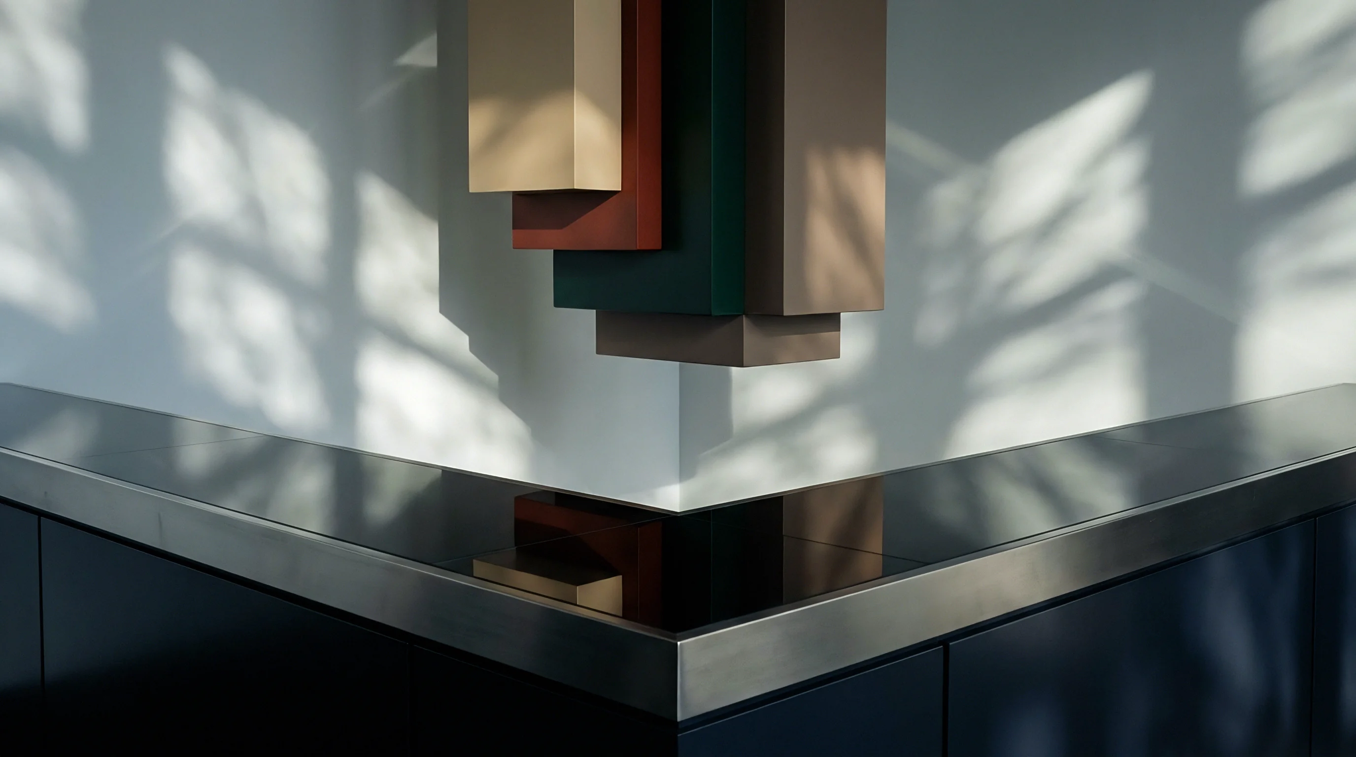 Layered rectangular blocks in diverse colors anchor a wall above a dark reflective metallic counter edge beneath diffused leaf shadows.